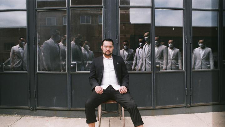 A man sits on a stool in front of a glass door reflecting multiple men in suits and face masks.