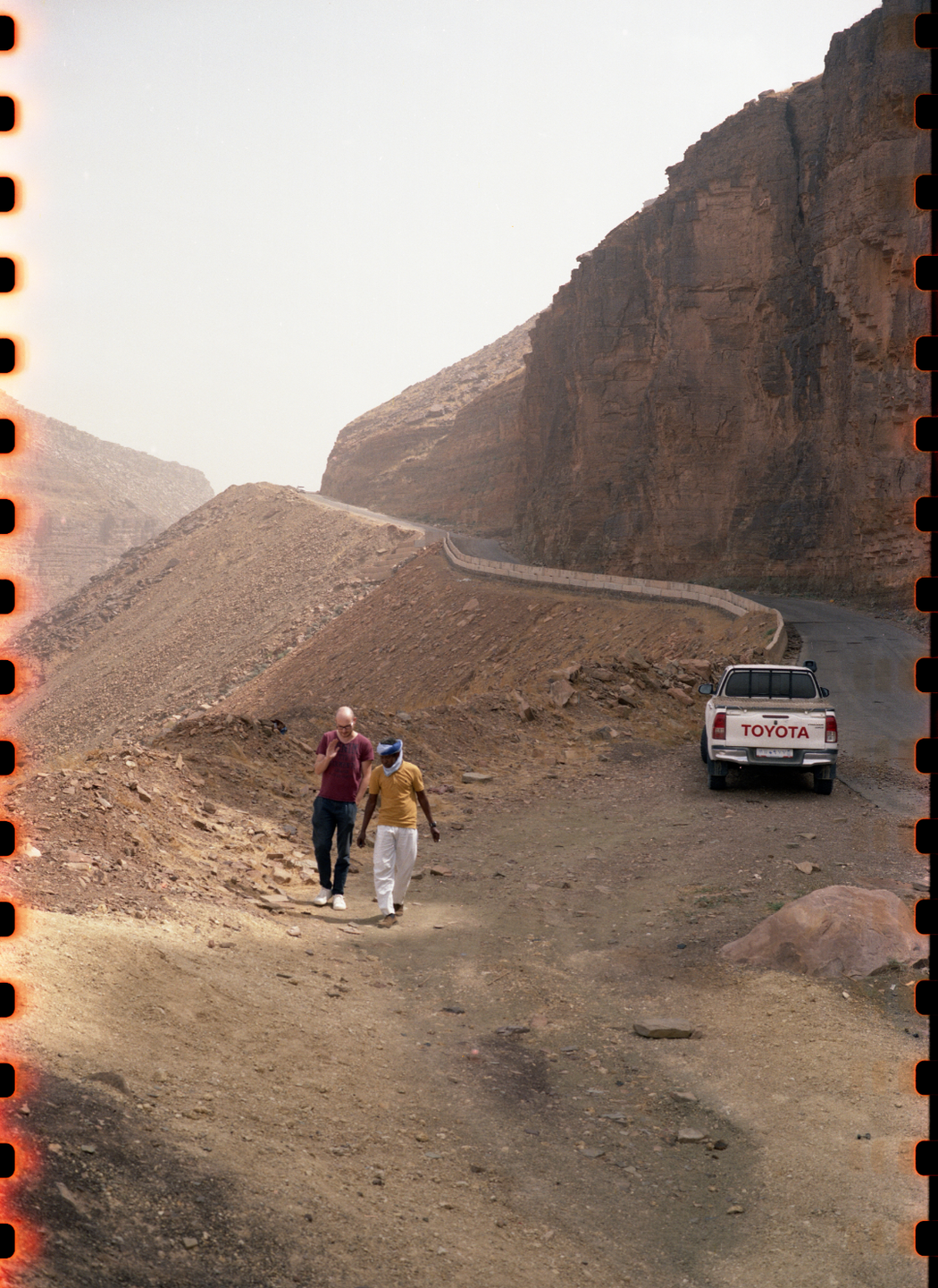 Two men walk along a dirt road next to a white pickup truck parked on the side of a winding mountain road.