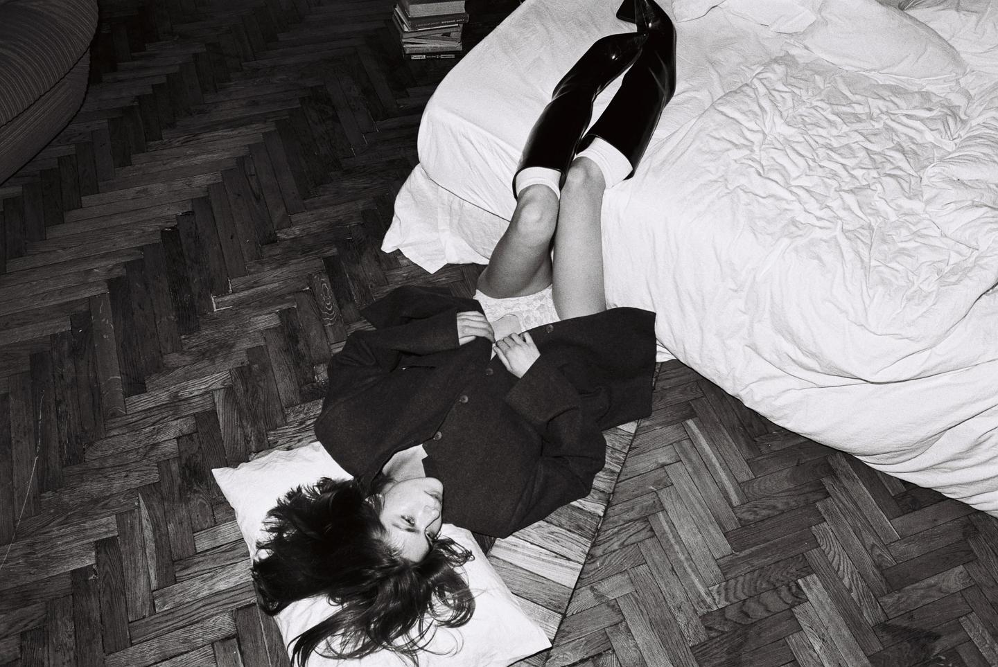 A woman lies on a pillow on a wooden floor next to a bed, wearing a coat and boots.