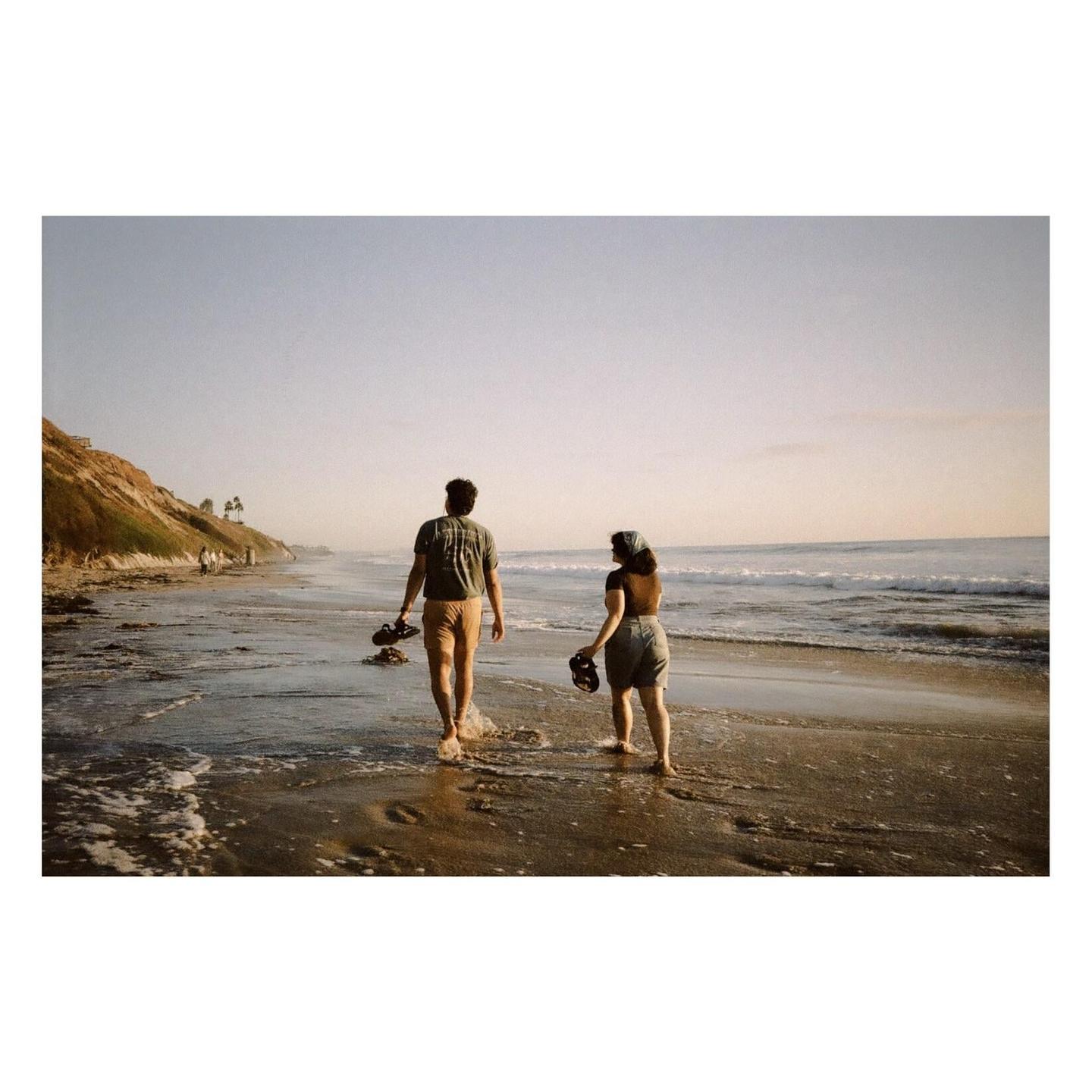 A couple walks barefoot along the water's edge on a beach with a cliff on the left.
