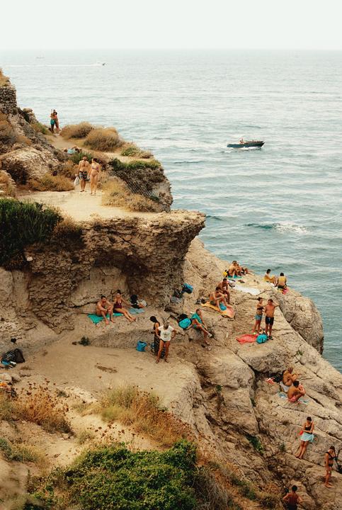 People are relaxing on a rocky cliff overlooking the ocean, with a boat visible in the distance.