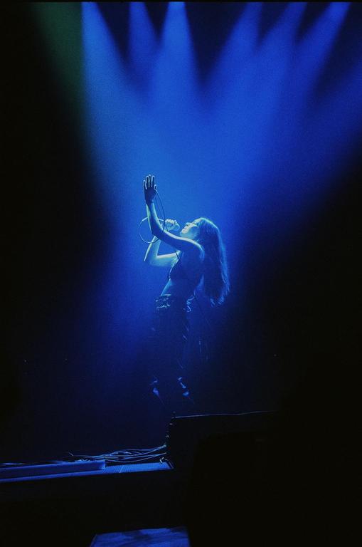 A woman sings into a microphone on a stage illuminated by blue spotlights.