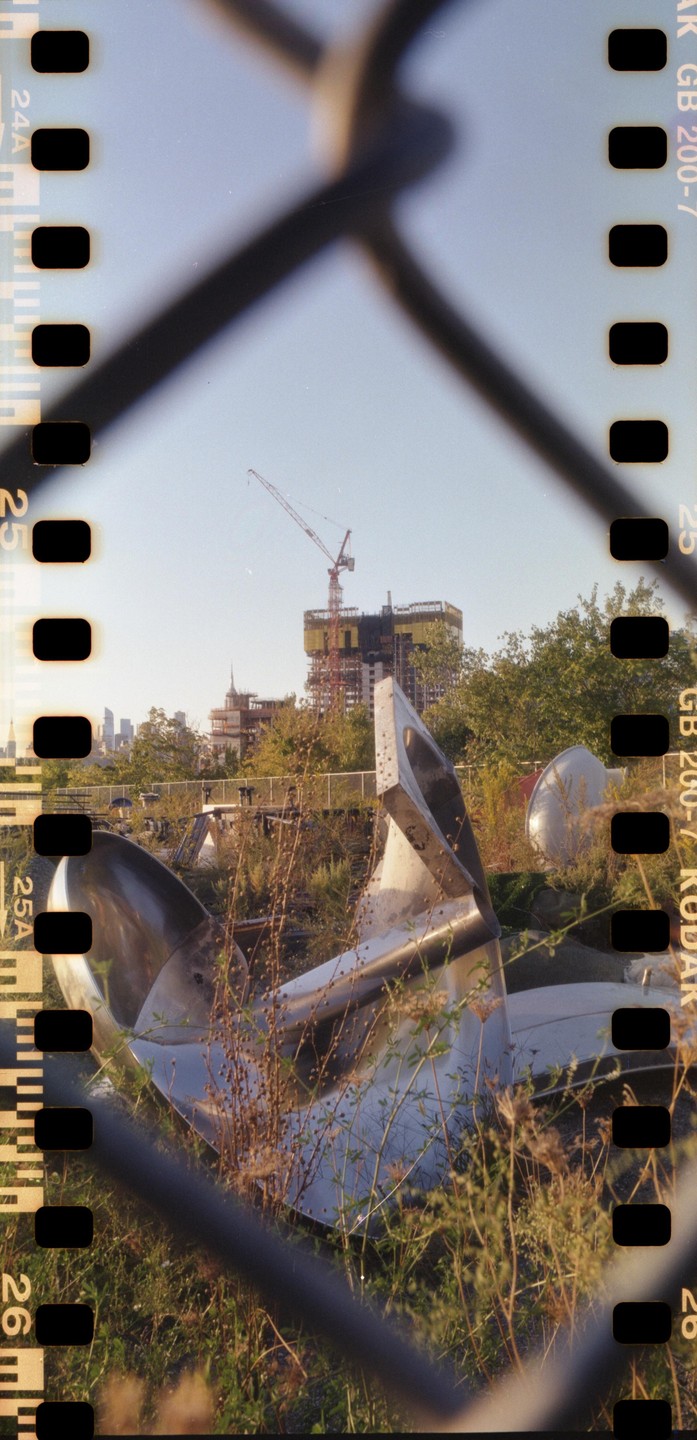 A metal sculpture is visible through a chain link fence, with a construction crane and city skyline in the background.