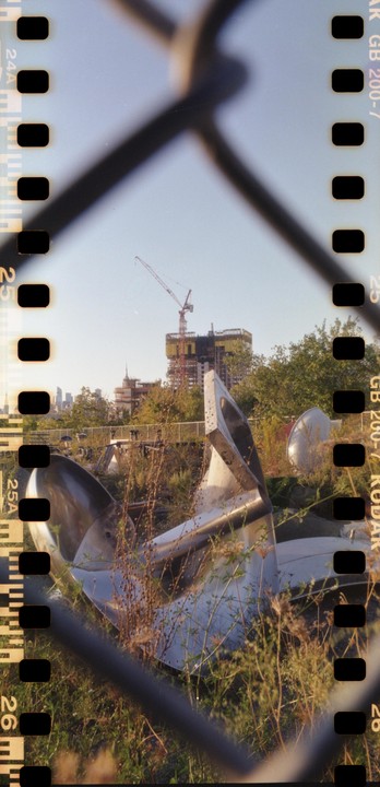 A metal sculpture is visible through a chain link fence, with a construction crane and city skyline in the background.