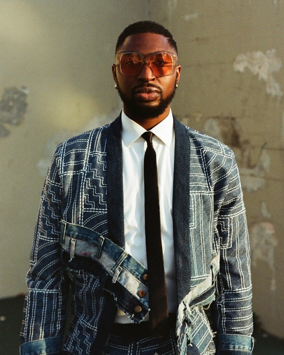 A man wearing a patterned denim jacket with deconstructed elements, a white shirt, a dark tie, and orange-tinted sunglasses poses for a portrait.