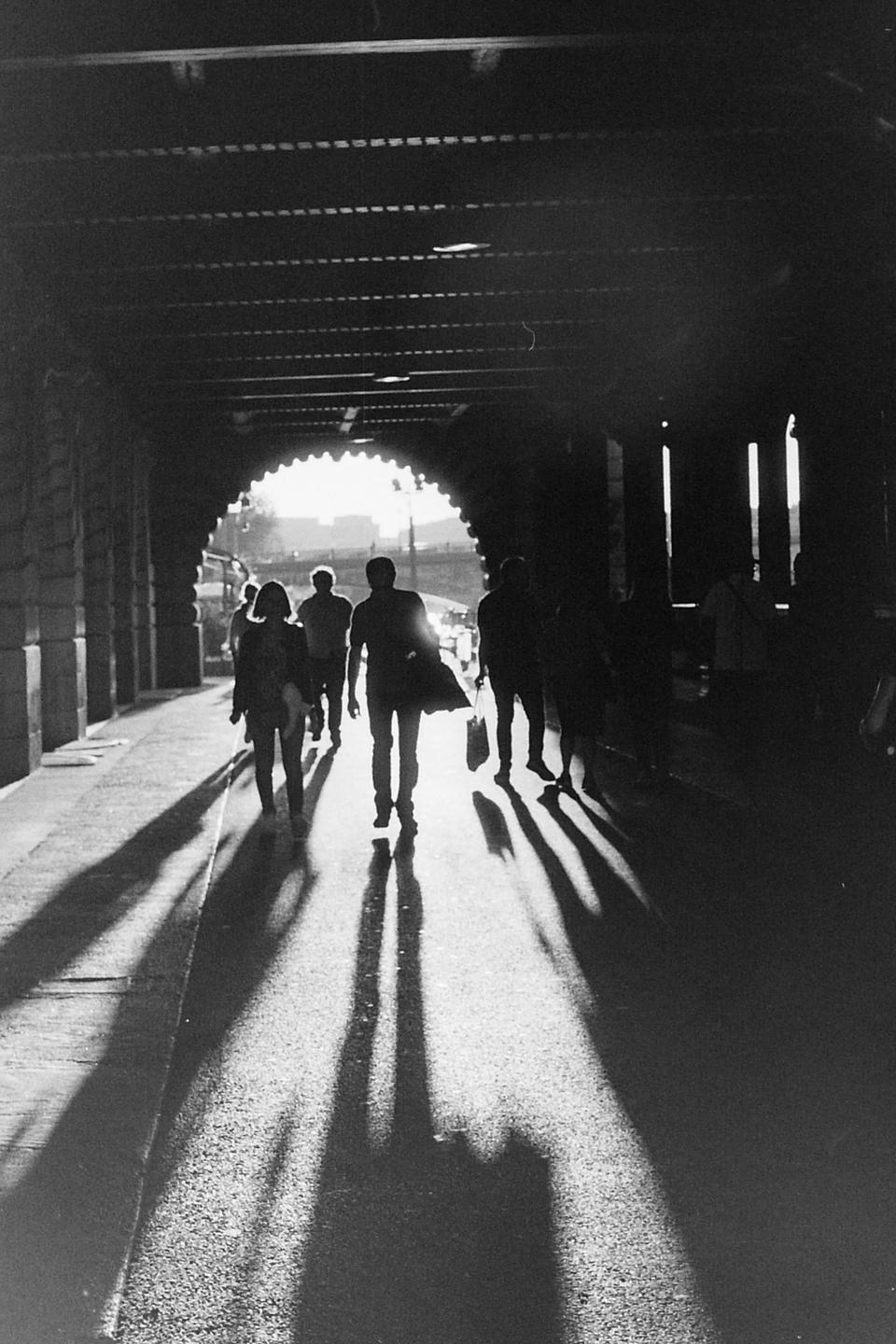 People walk through a dark tunnel with bright light at the end, casting long shadows on the ground.
