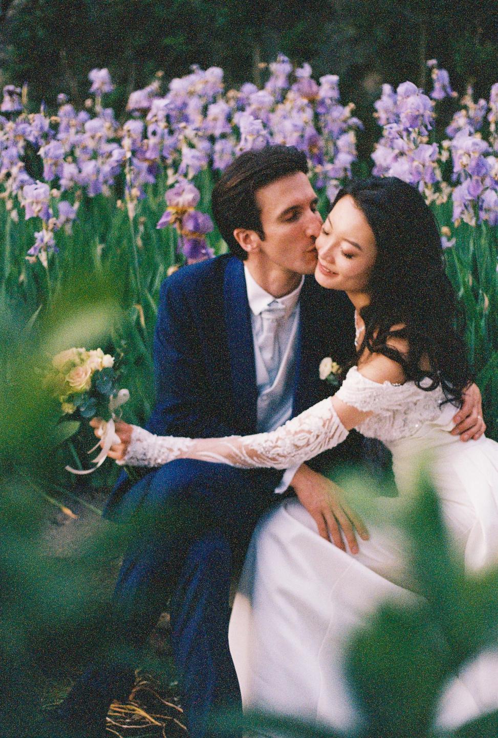 A man in a suit kisses a woman in a wedding dress on the cheek while sitting in a field of purple flowers.