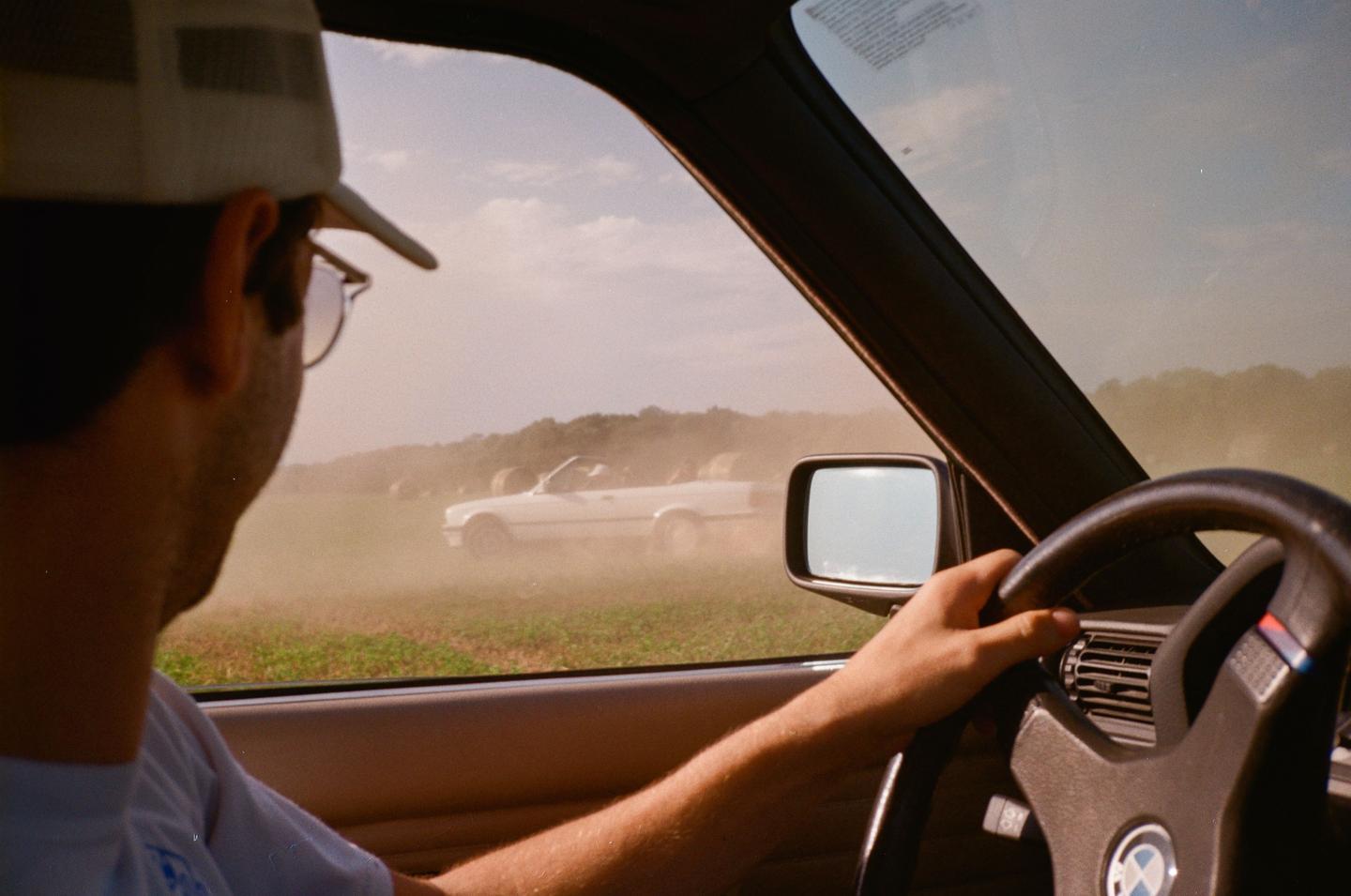 A man in a car looks out the window at another car driving through a dusty field.