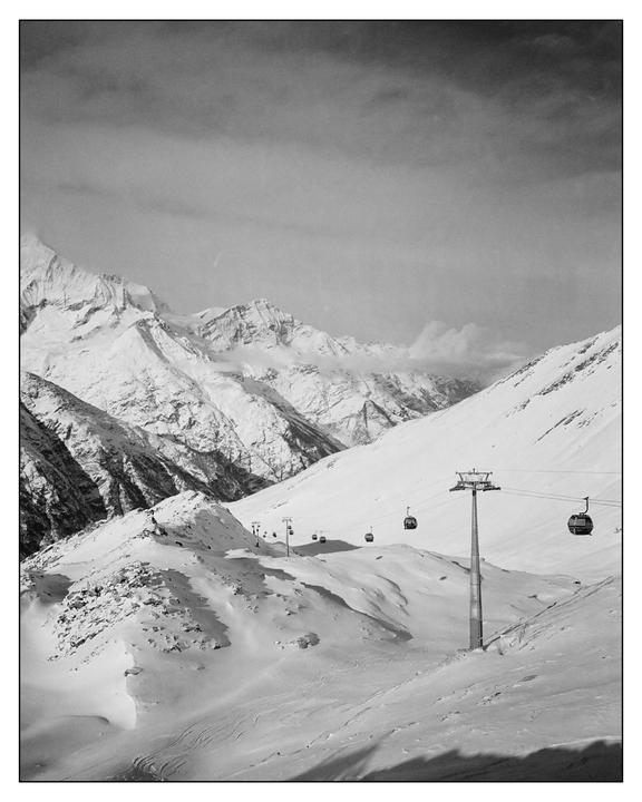A ski lift with several gondolas ascends a snow-covered mountain range under a cloudy sky.