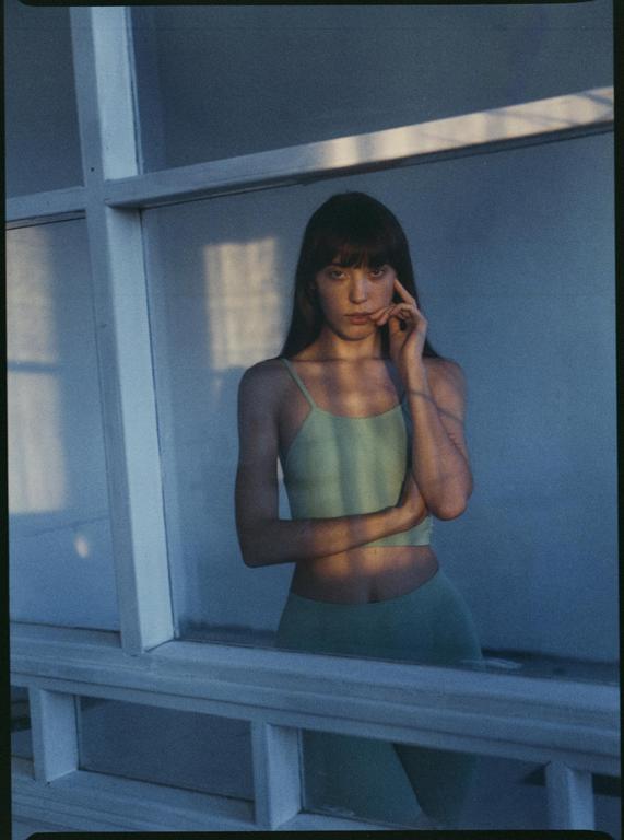 A woman in a light green top and pants stands behind a window with shadows cast across her body.