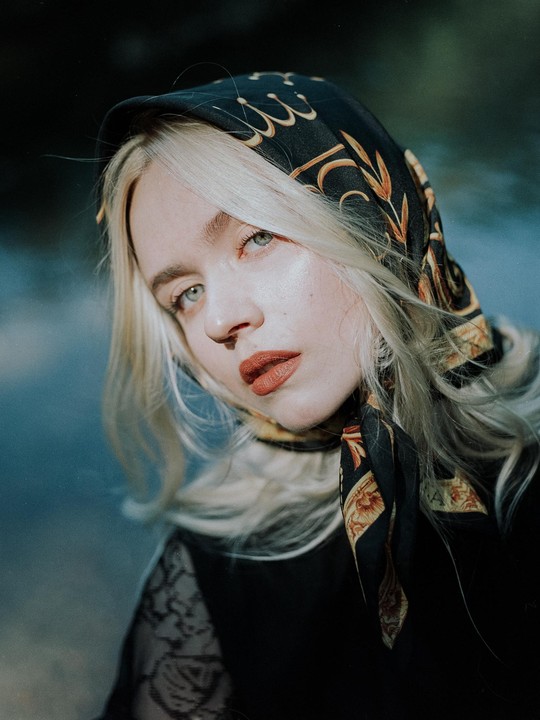 A woman with blonde hair and a patterned headscarf looks towards the viewer.