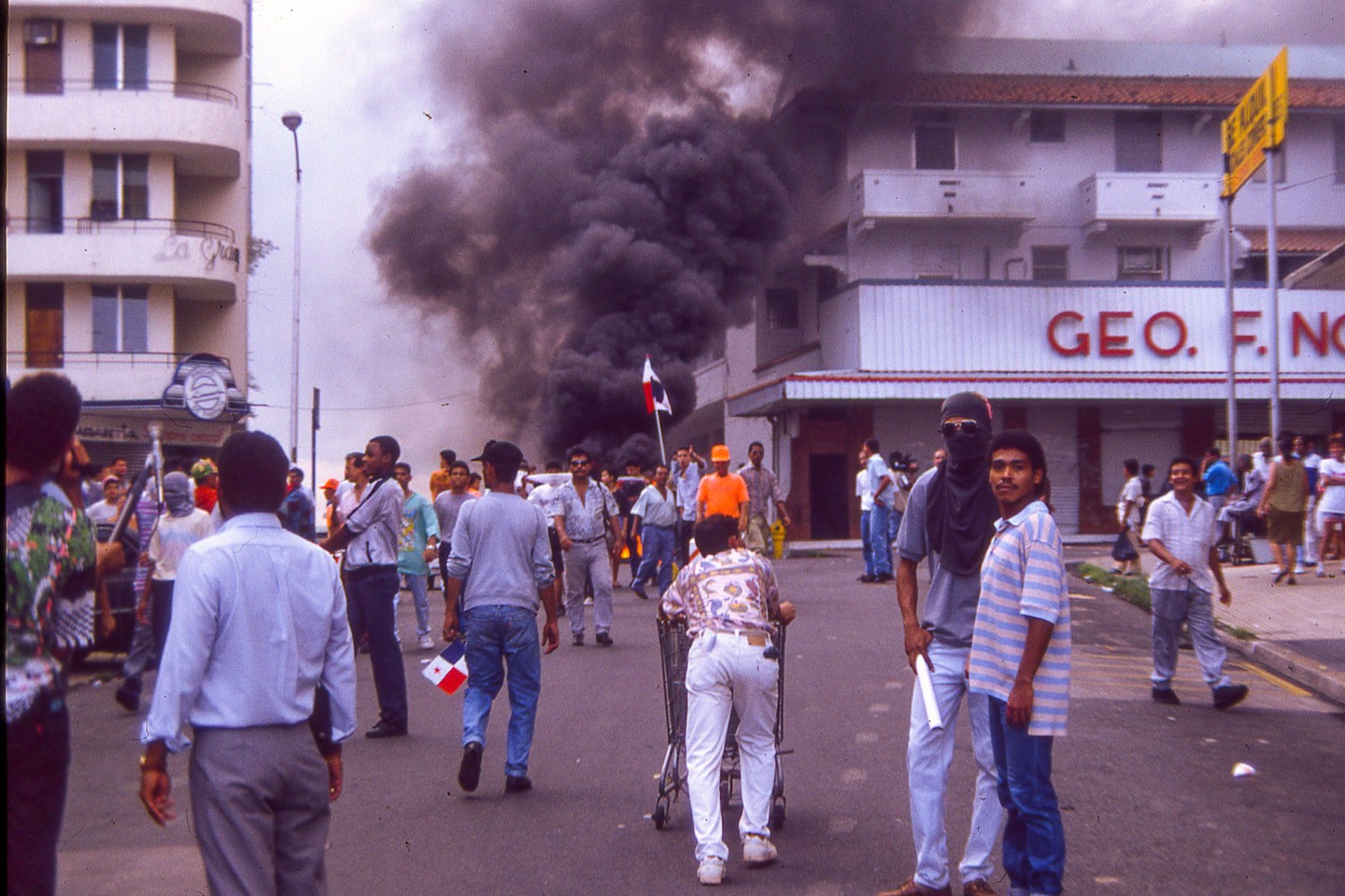 A crowd of people are gathered on a street with a large plume of dark smoke rising in the background.