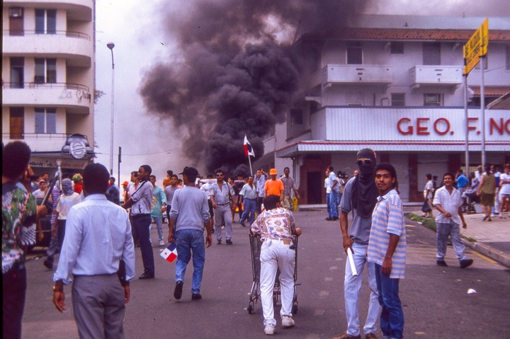 A crowd of people are gathered on a street with a large plume of dark smoke rising in the background.