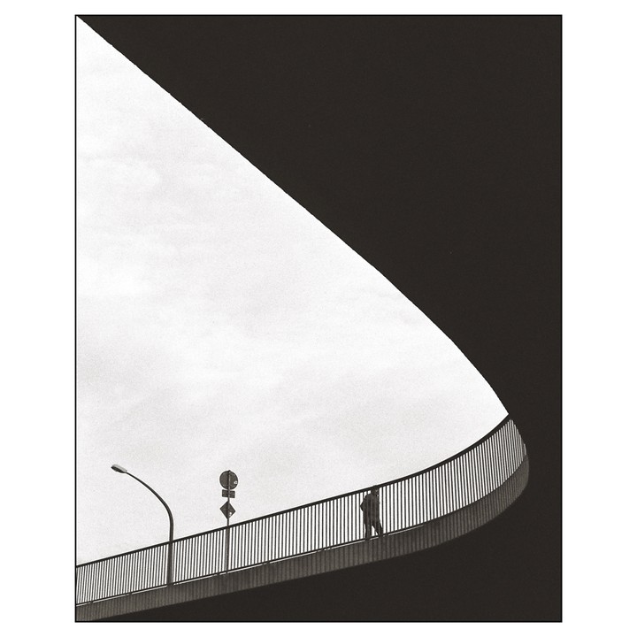 A person walks on a curved bridge with a street light and signs against a bright sky.