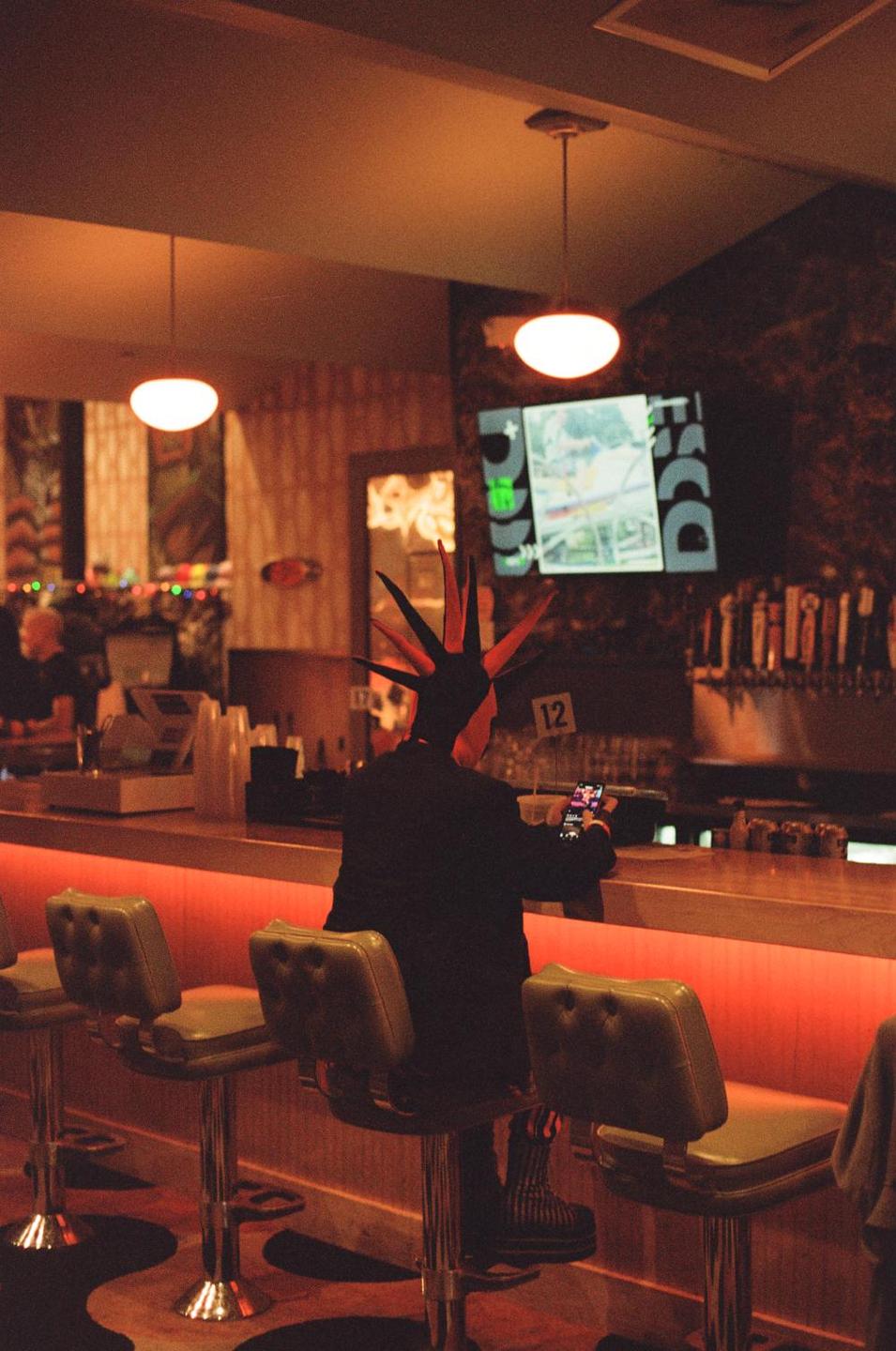 A person with a mohawk sits at a bar counter, looking at a phone.