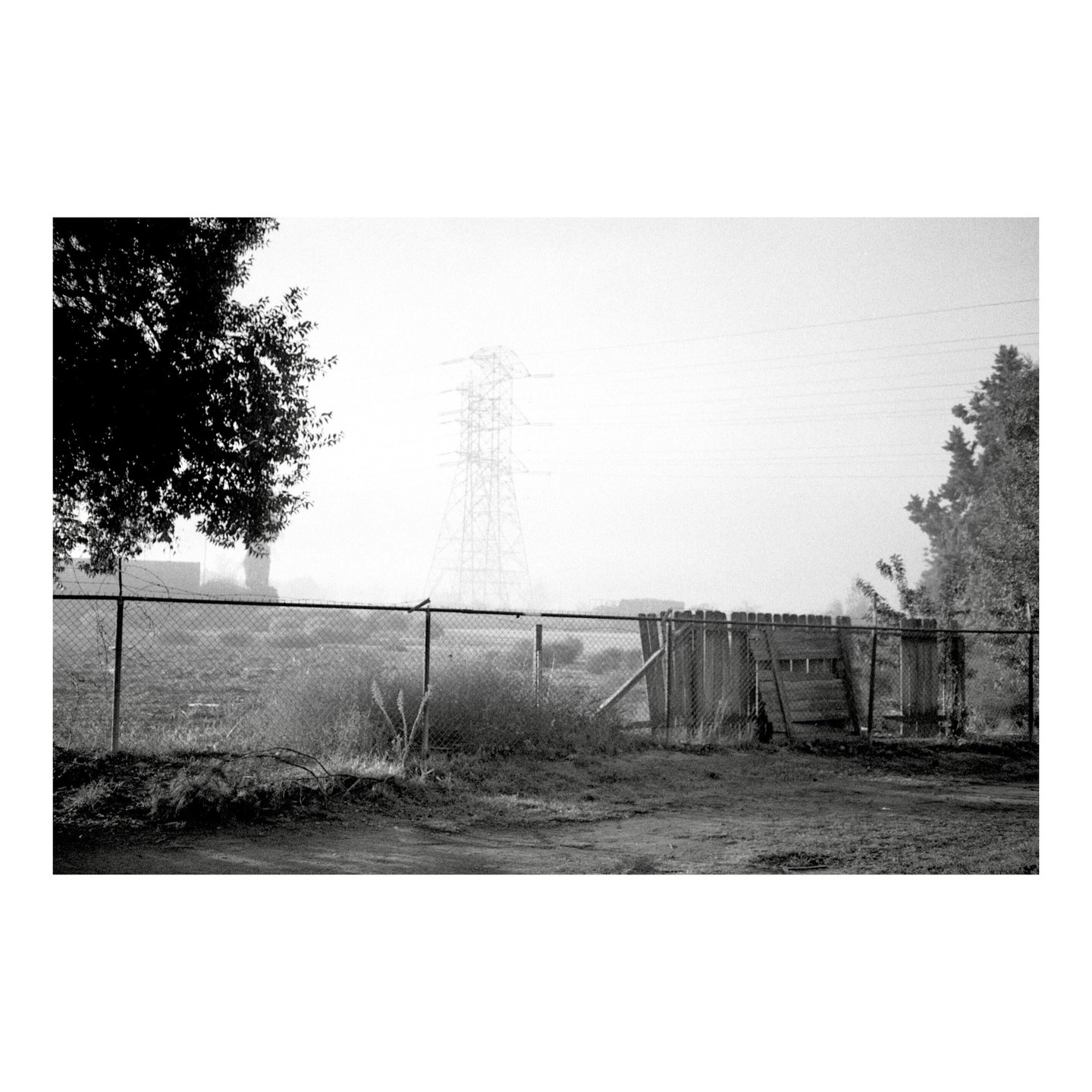 A chain link fence and a wooden fence stand in a field with a large power line tower in the distance on a foggy morning.