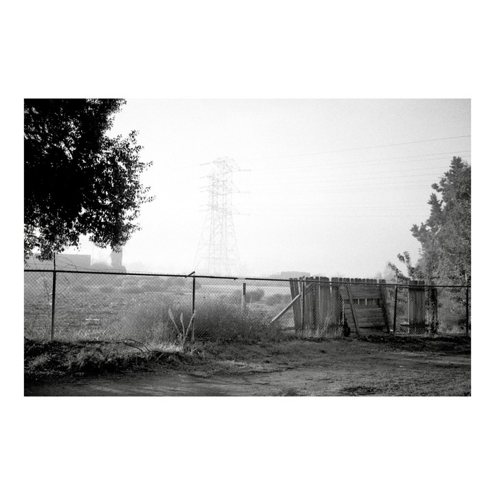 A chain link fence and a wooden fence stand in a field with a large power line tower in the distance on a foggy morning.