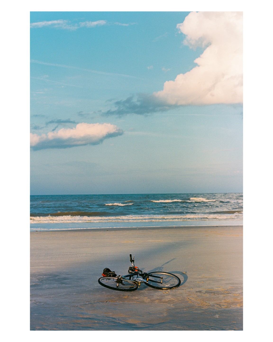 A bicycle lies on its side on a sandy beach with ocean waves in the background under a blue sky with clouds.