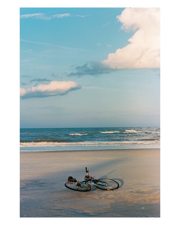 A bicycle lies on its side on a sandy beach with ocean waves in the background under a blue sky with clouds.