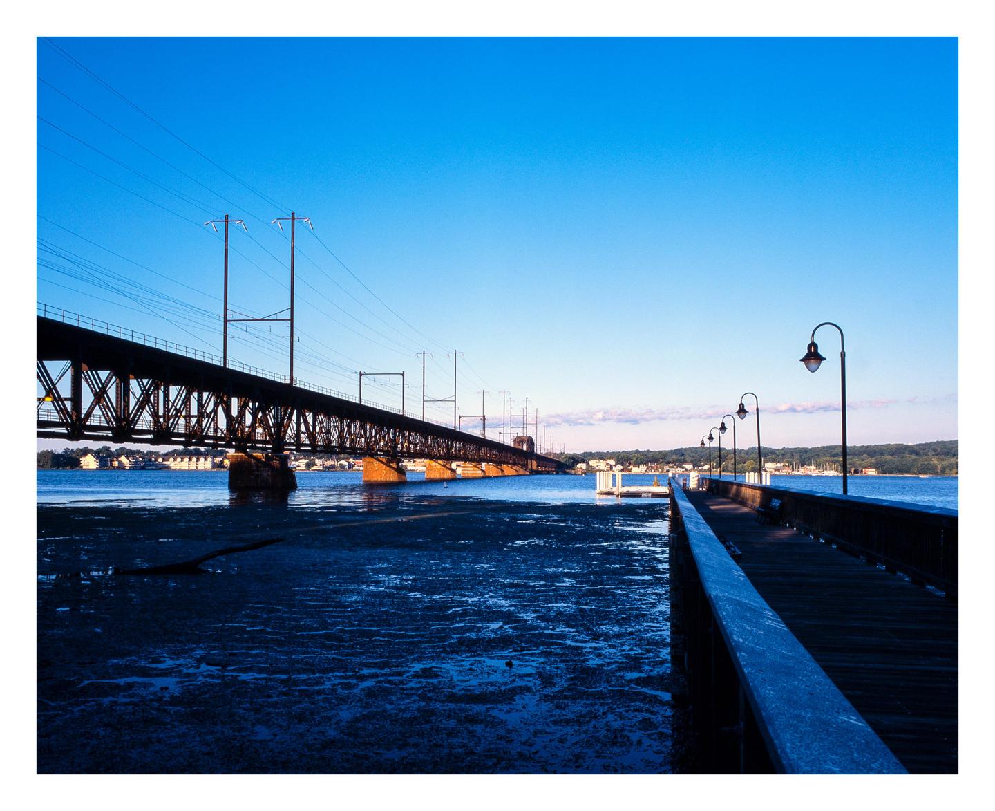 A long pier with streetlights extends into a body of water next to a large bridge under a clear sky.