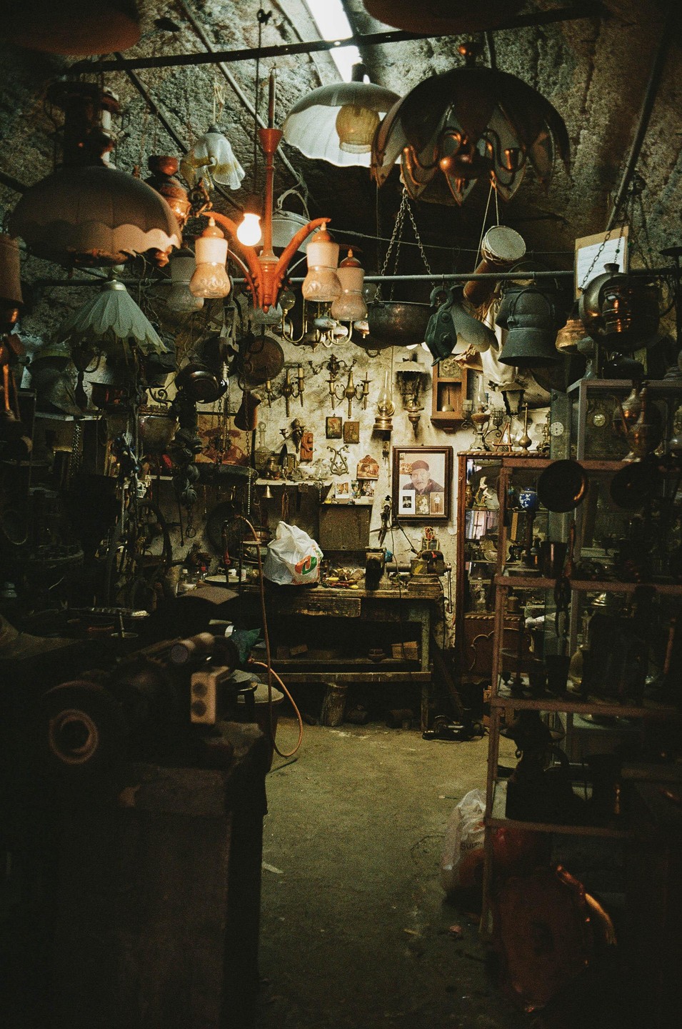 A cluttered shop is filled with various lamps, chandeliers, and other items, with a framed portrait of a man on the back wall.
