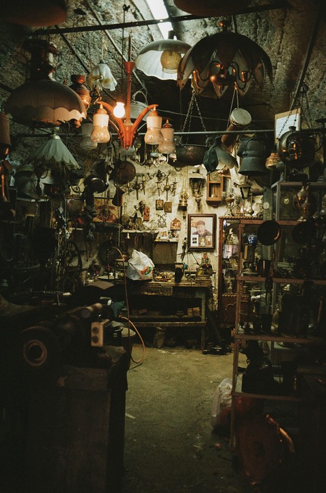 A cluttered shop is filled with various lamps, chandeliers, and other items, with a framed portrait of a man on the back wall.