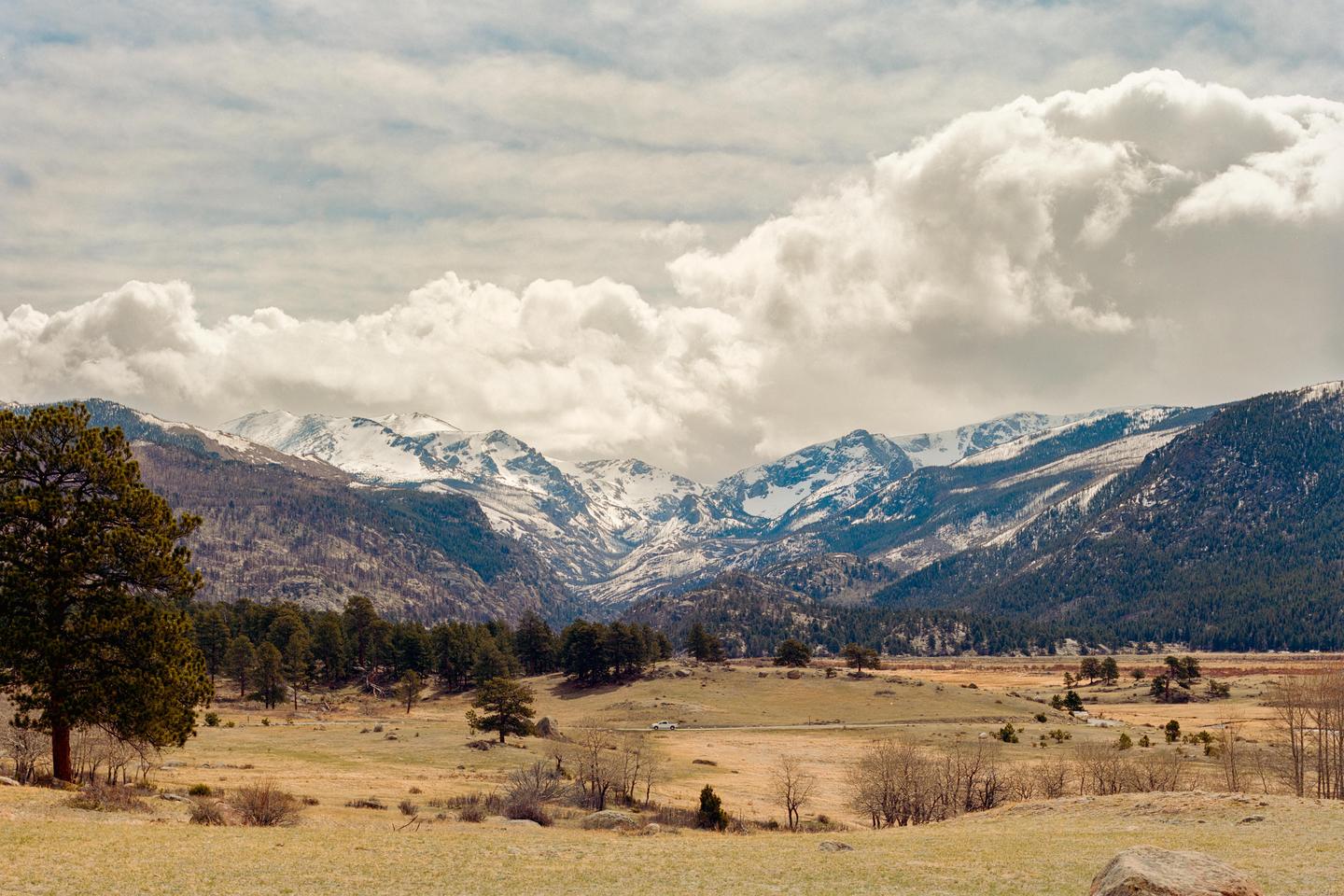 A vast landscape shows snow-capped mountains under a cloudy sky, with a valley of trees and fields in the foreground.