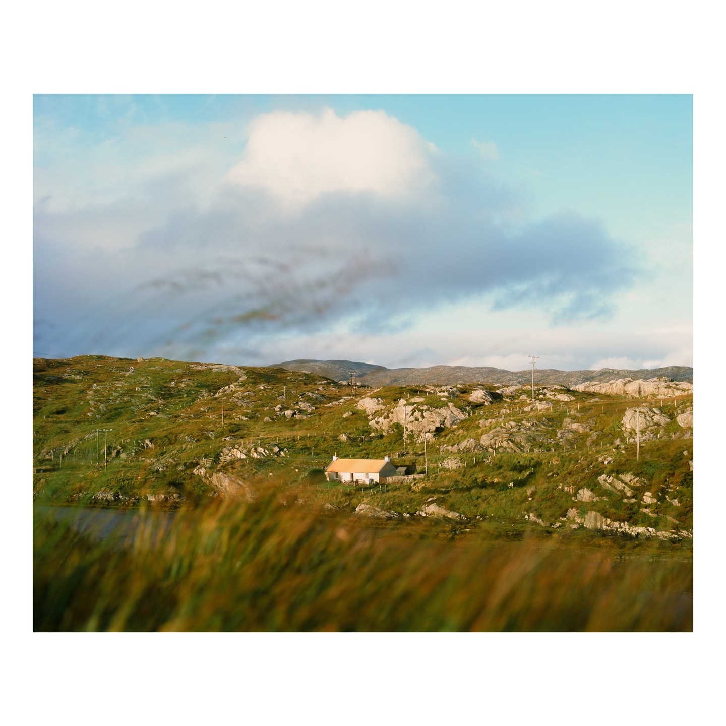 A small white house with a yellow roof sits in a green, rocky landscape under a cloudy sky.