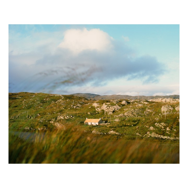 A small white house with a yellow roof sits in a green, rocky landscape under a cloudy sky.