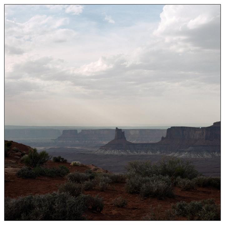 A vast canyon landscape with rock formations and sparse vegetation is bathed in sunlight under a cloudy sky.