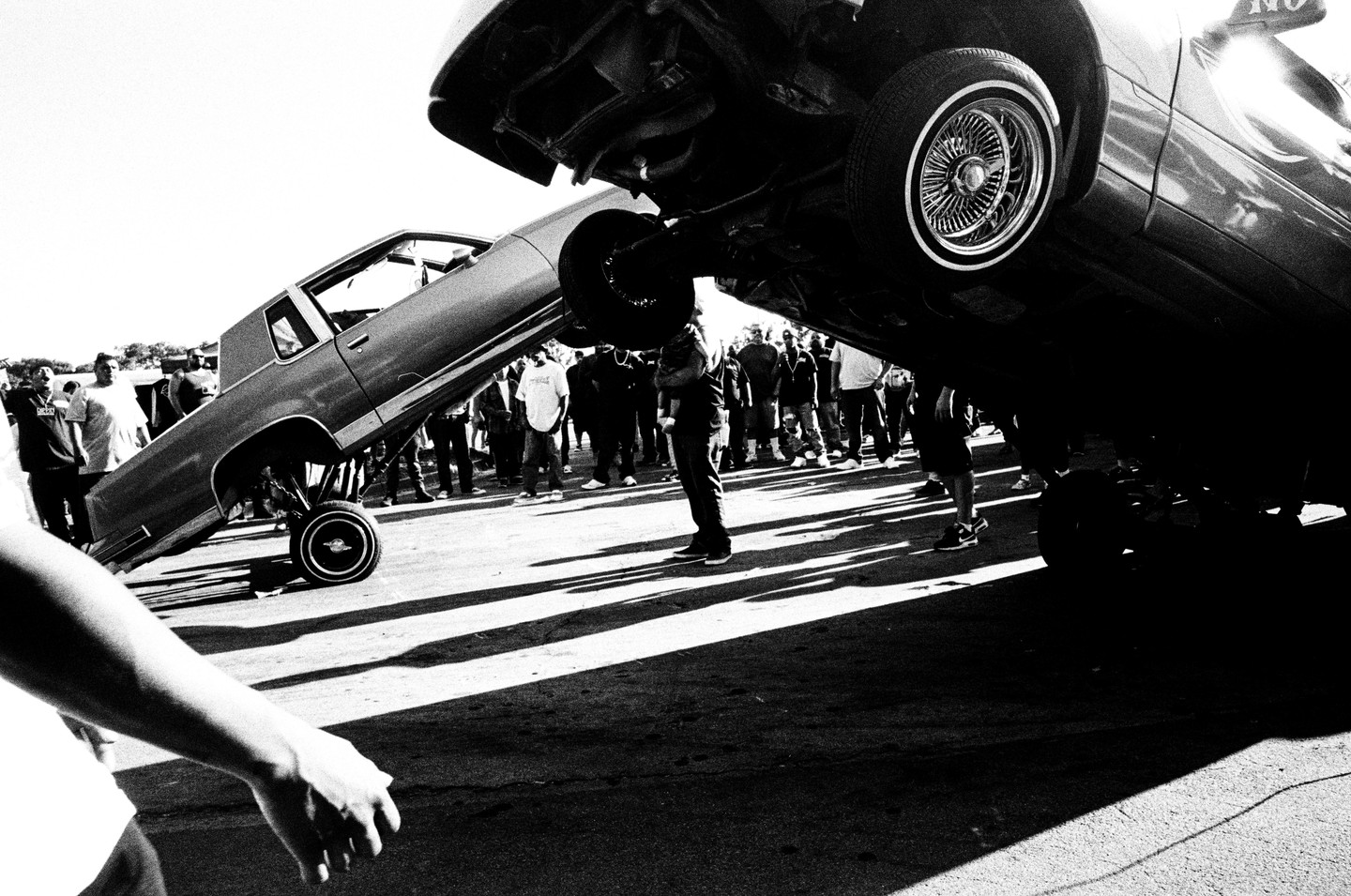 Two lowrider cars are bouncing on their hydraulic suspensions in front of a crowd of people.