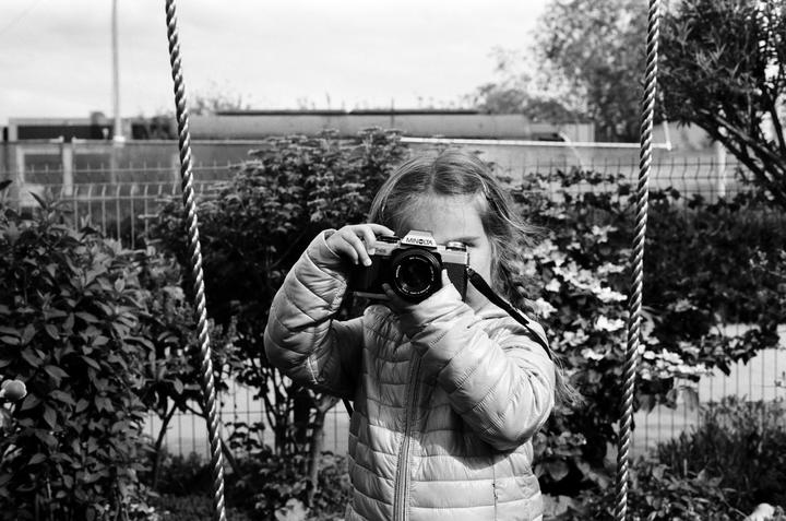 A young girl holds a camera up to her eye, framed by ropes on either side, with bushes and a fence in the background.