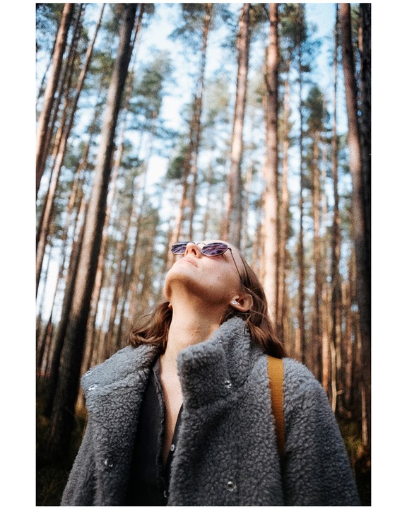A woman in a grey coat and sunglasses looks up at tall trees in a forest.