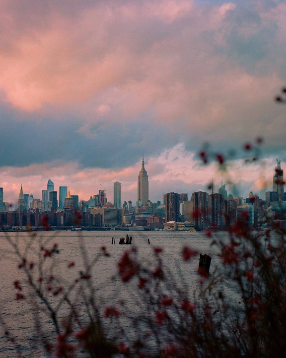 The New York City skyline is visible across the water at sunset, with blurred red foliage in the foreground.