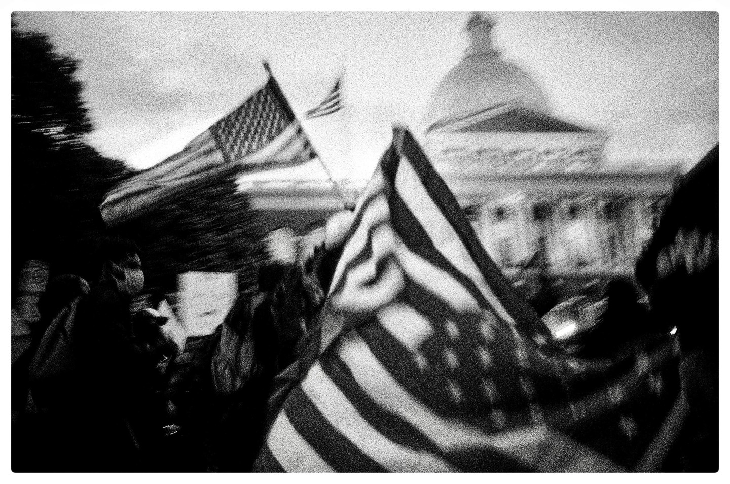 Two American flags wave in front of a domed building with people in the foreground.