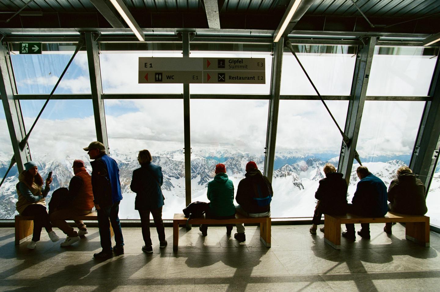 Several people are looking out of a large window at a snowy mountain range.