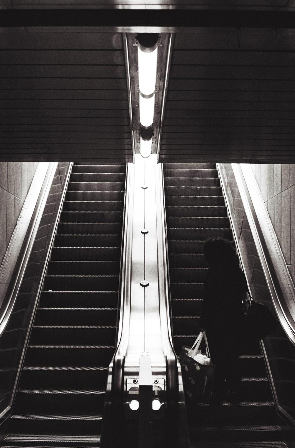 A person stands on an escalator next to another escalator going in the opposite direction, with a bright light fixture above.