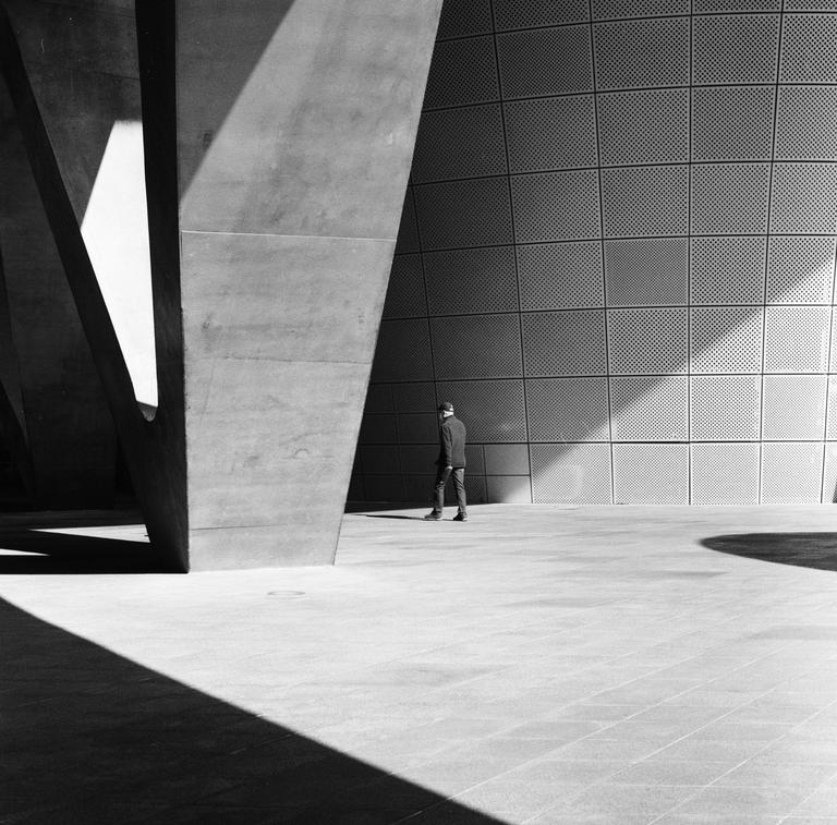 A man walks on a paved surface next to a large concrete structure and a textured wall with geometric patterns of light and shadow.