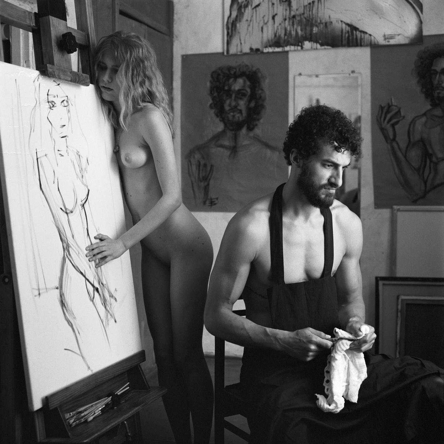 A nude woman leans against an easel with a charcoal drawing of a female figure while a man in an apron sits nearby.