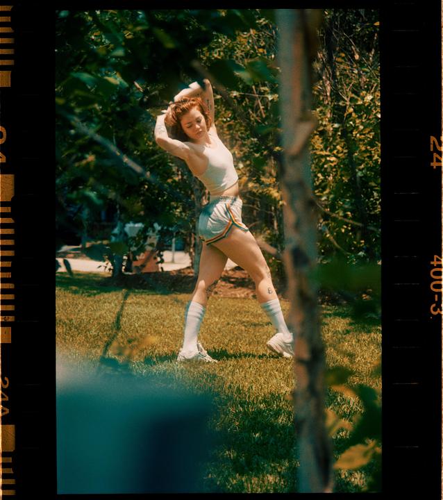 A woman with red hair stretches her arms above her head in a grassy area surrounded by trees.