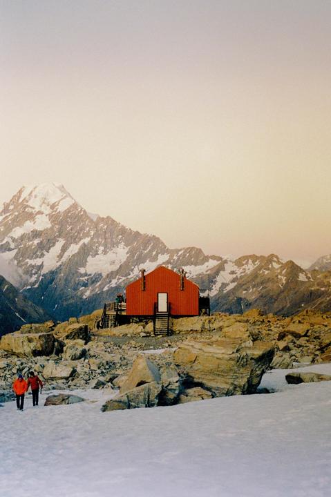 Two people walk on a snowy mountain path towards a red cabin with a snow-capped mountain in the background.
