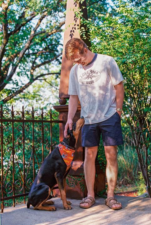 A man wearing a t-shirt and shorts pets a doberman dog wearing a bandana next to a metal fence and a stone pillar.
