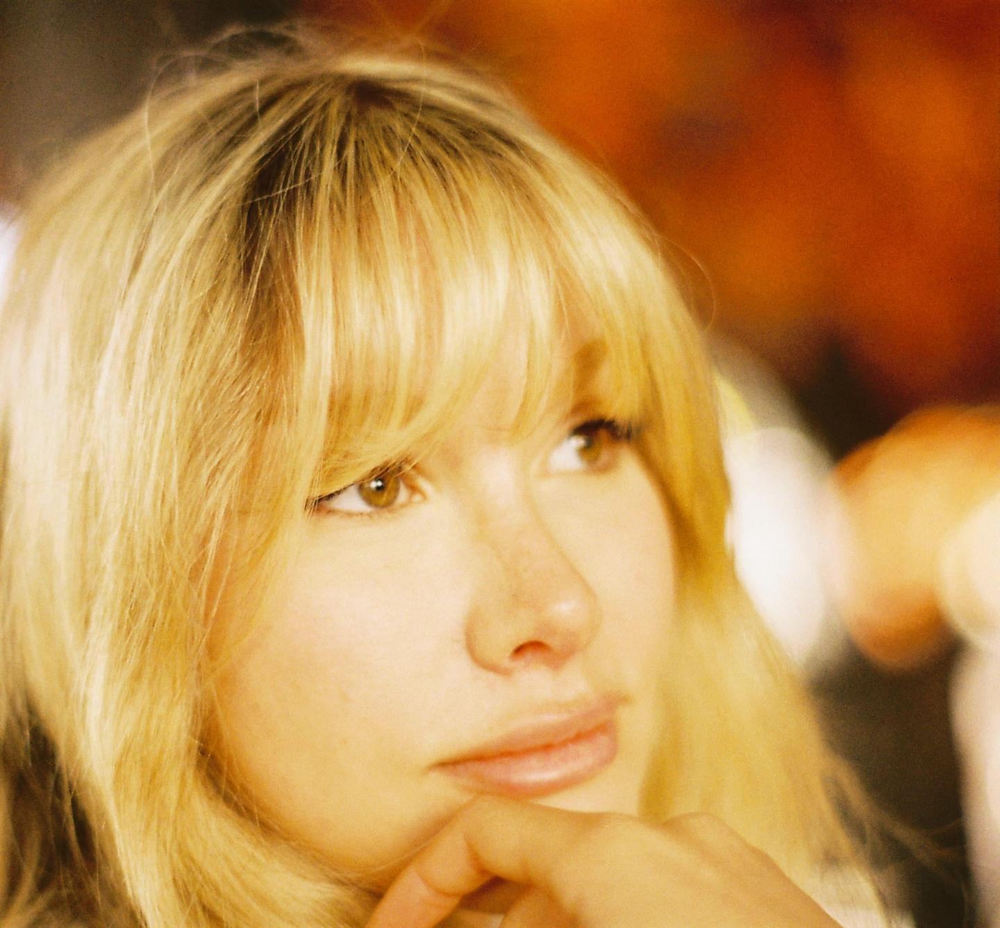 A woman with blonde hair and bangs looks to the side with her hand on her chin.