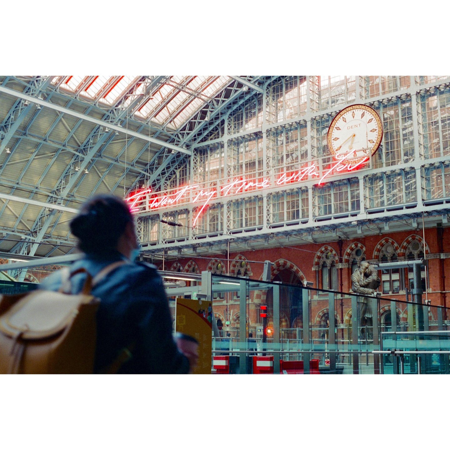 A woman with a backpack stands in a large train station with a neon sign and a clock.