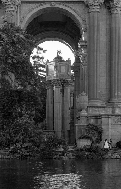 A couple stands by a body of water in front of a large classical building with arches and columns.