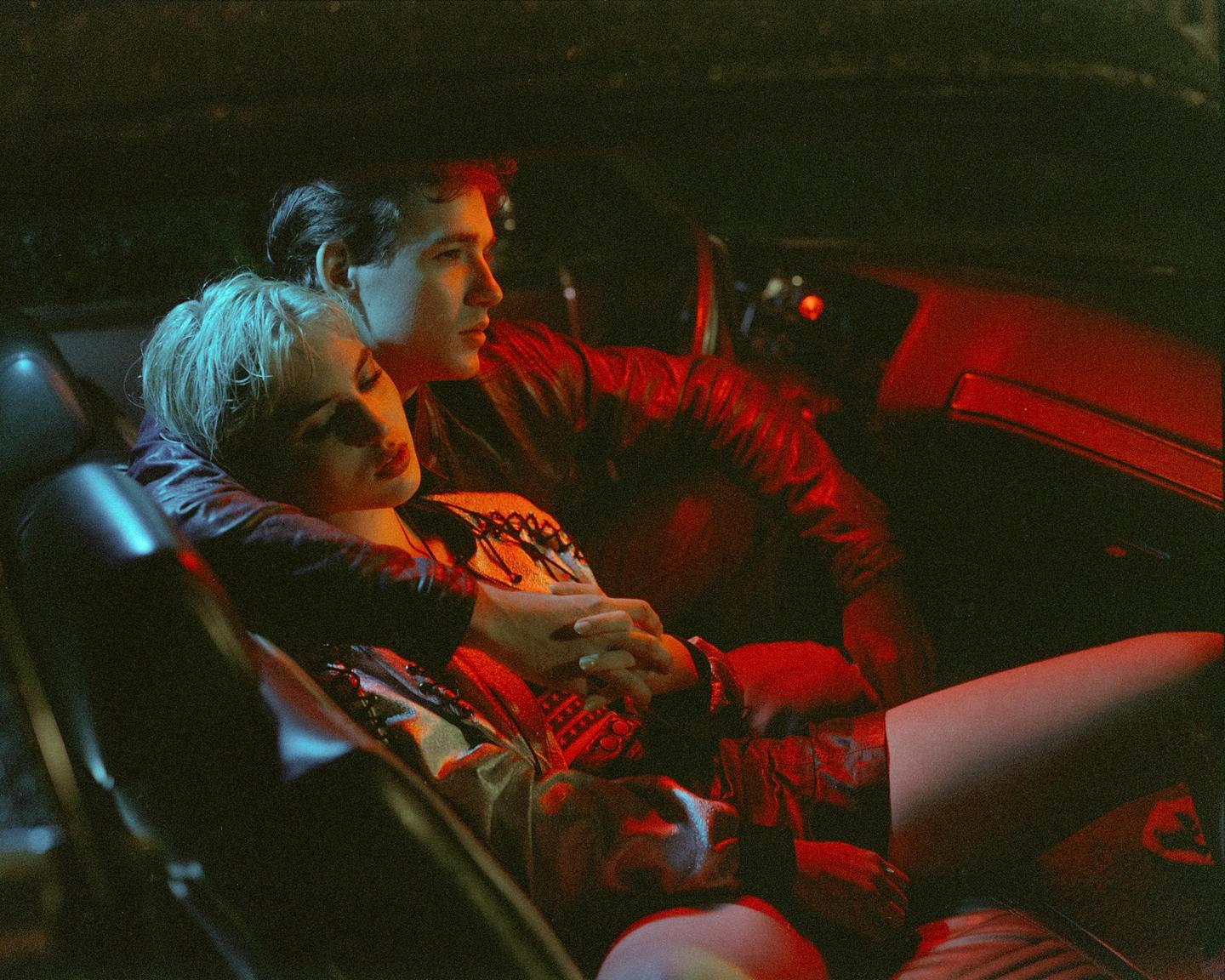 A man and a woman are sitting in a car at night, illuminated by red and blue light.