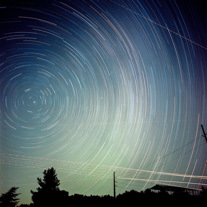 Star trails arc across the night sky above a silhouetted landscape with trees and power lines.