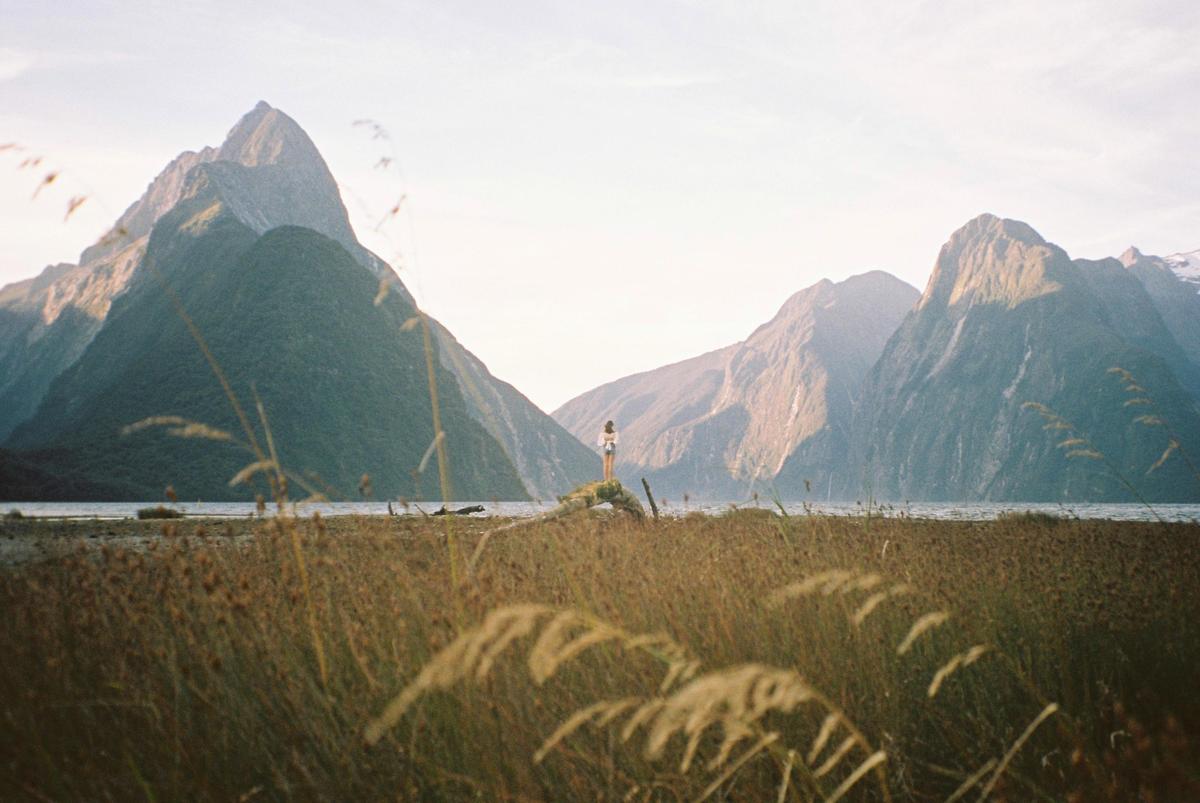 A woman stands on a rock in a field of tall grass, looking out at a body of water surrounded by mountains.
