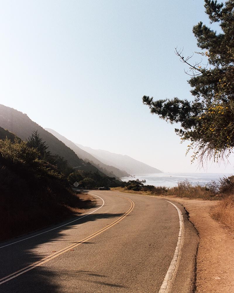 A winding road follows a coastline with mountains in the background.