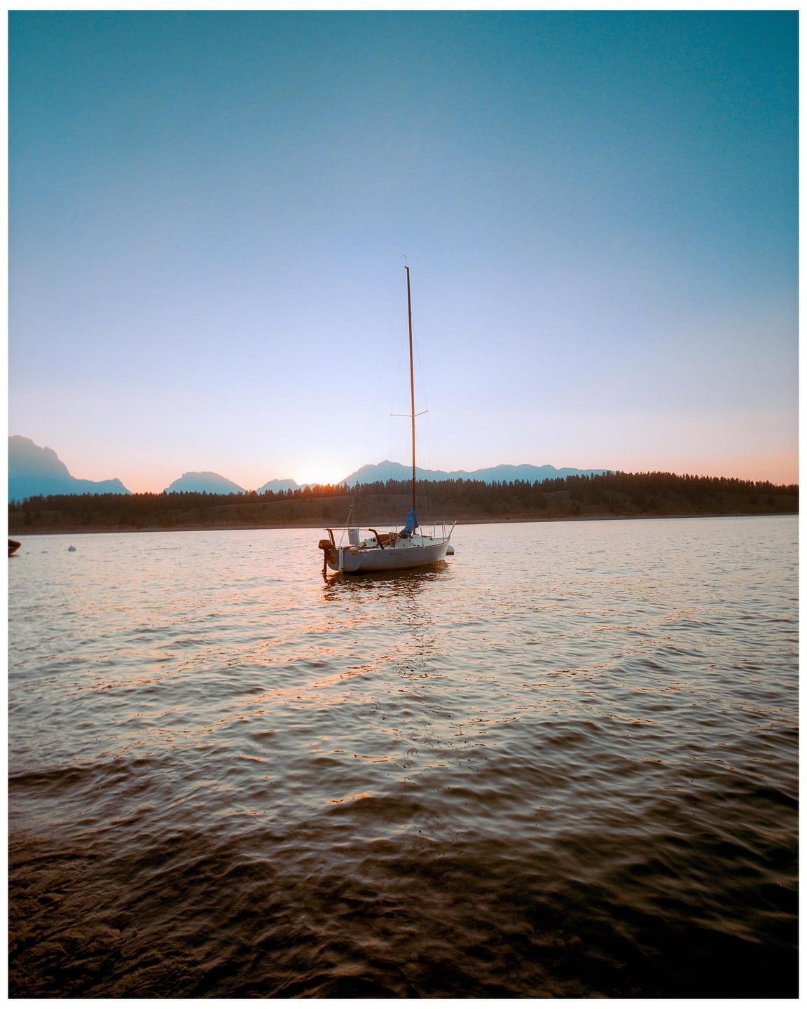 A sailboat floats on a lake with mountains and a forest in the background at sunset.